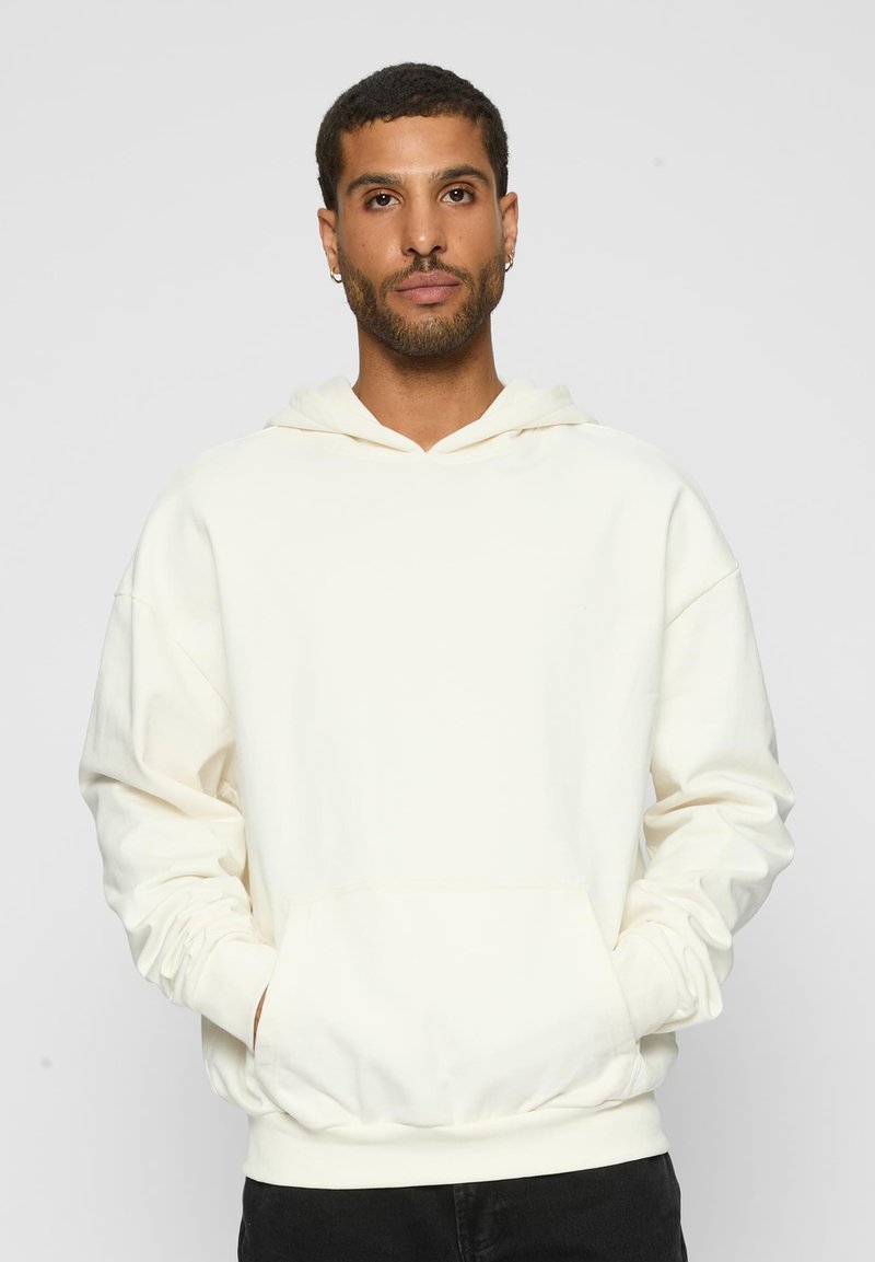Felpa crema realizzata in tessuto morbido, con tasca frontale, cappuccio con coulisse, vestibilità comoda e polsini a costine. Indossata sopra pantaloni neri.
