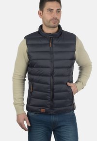 Dunkelblaues Steppweste mit hohem Kragen, horizontalem Steppmuster und Reißverschlussfront. Verfügt über Lederakzente und seitliche Taschen. Über einem leichten Pullover getragen.
