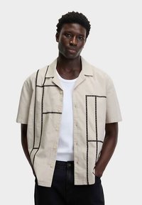 Beiges Kurzarmhemd mit lockerem Schnitt, das dunkle Linienmuster über den Stoff hat und vorne mit Knöpfen versehen ist. Über einem weißen Hemd getragen.