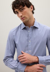 Camisa de botones con rayas verticales en azul y blanco, cuello y mangas largas. Tejido suave, corte ajustado, con un detalle de un solo bolsillo.