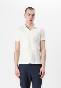 Polo tričko s krátkým rukávem, ribbed pletenina v krémové barvě, s výstřihem do V a kontrastním límcem. Spárováno s tmavě modrými kalhotami.
