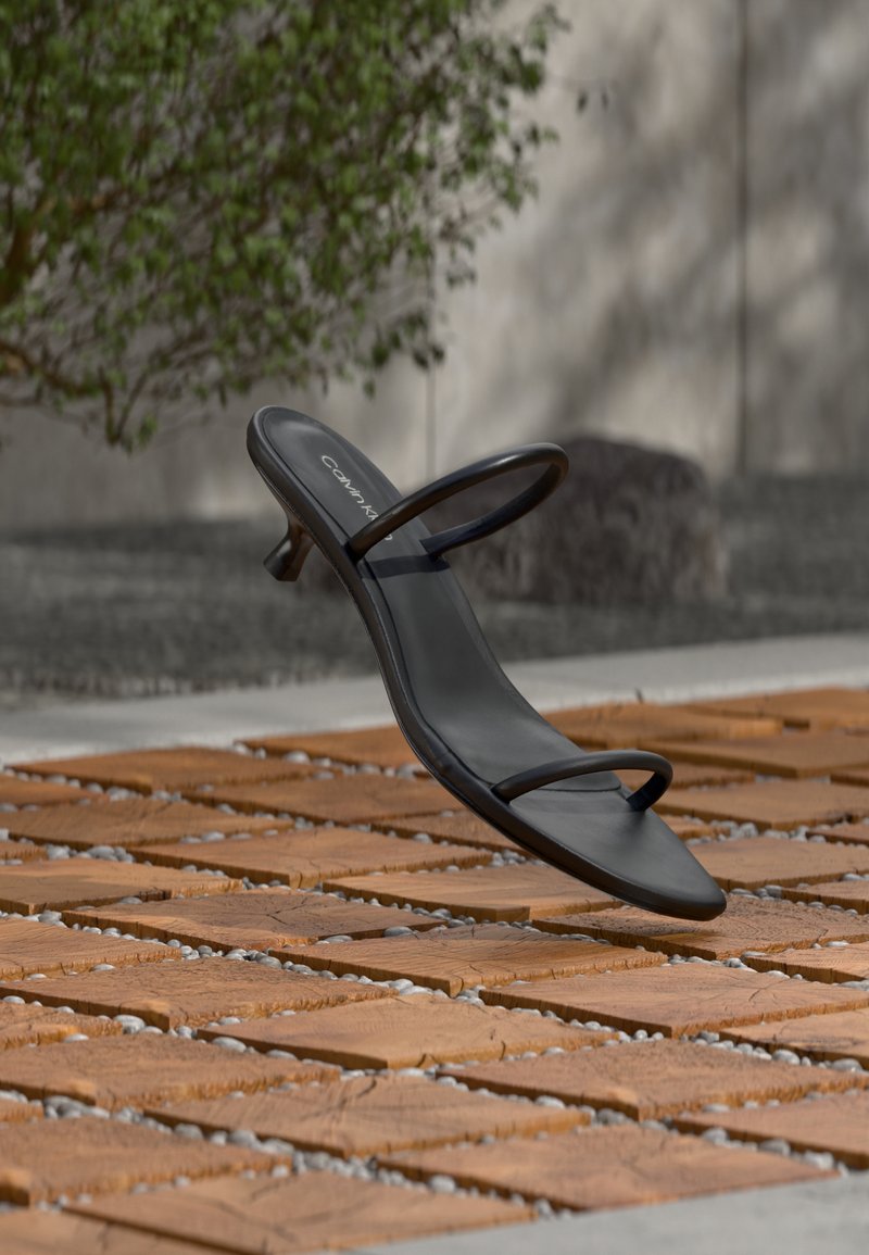 Μαύρο ψηλοτάκουνο σανδάλι Calvin Klein με λεπτές λουρίδες, ισορροπημένο σε ξύλινες πλάκες έξω κοντά σε έναν τοίχο και φυλλώματα.