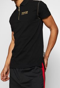 Zwarte polo met korte mouwen, ritskraag, gouden borduurwerk "VERSACE JEANS COUTURE," en contrasterende schouderstiksels.