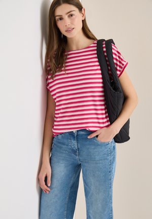 Jeune femme portant une chemise à rayures roses et blanches, un jean bleu et un sac à bandoulière noir, debout devant un mur beige.