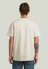 T-shirt di cotone beige con maniche corte, taglio dritto e scollatura a girocollo. Presenta una texture liscia e tatuaggi visibili sulla schiena.
