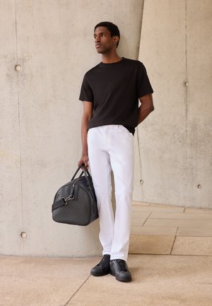 Hombre con camiseta negra y pantalones blancos está apoyado en una pared de cemento sosteniendo una bolsa de viaje de color gris oscuro con diseño en su mano izquierda.