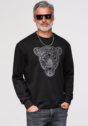 Mann mit grauem Haar und Bart, trägt schwarze Sonnenbrille, eine Ketten-Halskette, schwarzen Pullover mit besticktem Tiergesicht und graue Jeans mit den Händen in den Taschen.