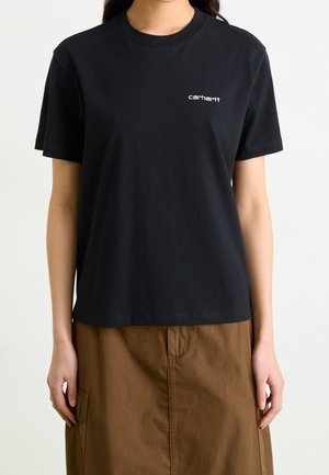 Kvinde iført en sort Carhartt T-shirt og brun nederdel, stående med armene afslappet langs siderne mod en ensartet lys baggrund.