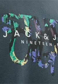 Tissu sombre avec des formes abstraites colorées formant de grandes lettres derrière un texte blanc disant « JACK & J NINETEEN M. »