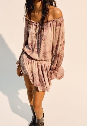 Femme portant une robe rose à motifs, épaules dénudées, manches longues, ourlet à volants et bottines noires, debout devant un fond uni.