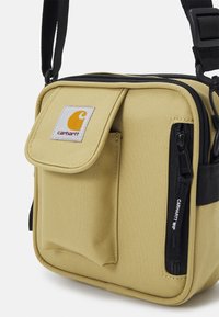 Żółta torba na ramię z płócienia z przednią kieszenią na klapę, boczną kieszenią na zamek i czarnym paskiem. Posiada logo Carhartt i trwałą tekstur�ę.