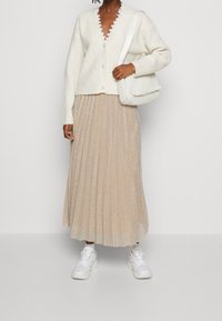 Biały kardigan z prążkowanego materiału o falbaniastym dekolcie, zestawiony z plisowaną, metaliczną, złotą spódnicą i białymi sneakersami; trzyma teksturowaną, białą torbę.