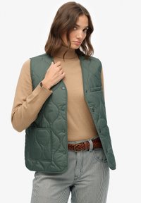 Zielona kamizelka pikowana z guzikami, boczną kieszenią i gładką teksturą; noszona na beżowym golfie i w paskowanych spodniach.