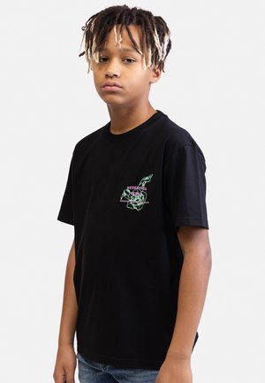 Chłopiec z jasnobrązowymi i ciemnobrązowymi dredami, ubrany w czarny t-shirt z nadrukiem i niebieskie jeansy, stoi na tle jednolitej białej ściany.