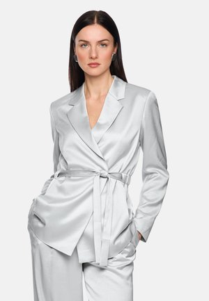 Frau mit langen dunklen Haaren, die ein silbernes Satin-Blazer mit Gürtel und passende Hosen trägt, Hände in den Taschen, vor hellem einfarbigem Hintergrund.