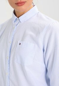 Pasiasta niebieska i biała koszula na guziki z kołnierzykiem, z kieszonką na piersi i subtelnym akcentem logo na kieszeni. Tekstura z bawełnianej tkaniny.