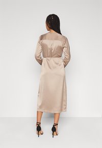 Satinė apvalkalo suknelė šviesiai beige spalvos, su ilgomis rankovėmis, prigludusia juosmeniu ir vidutinio ilgio iki blauzdų. Suderinta su juodais diržais aukštais kulnais.