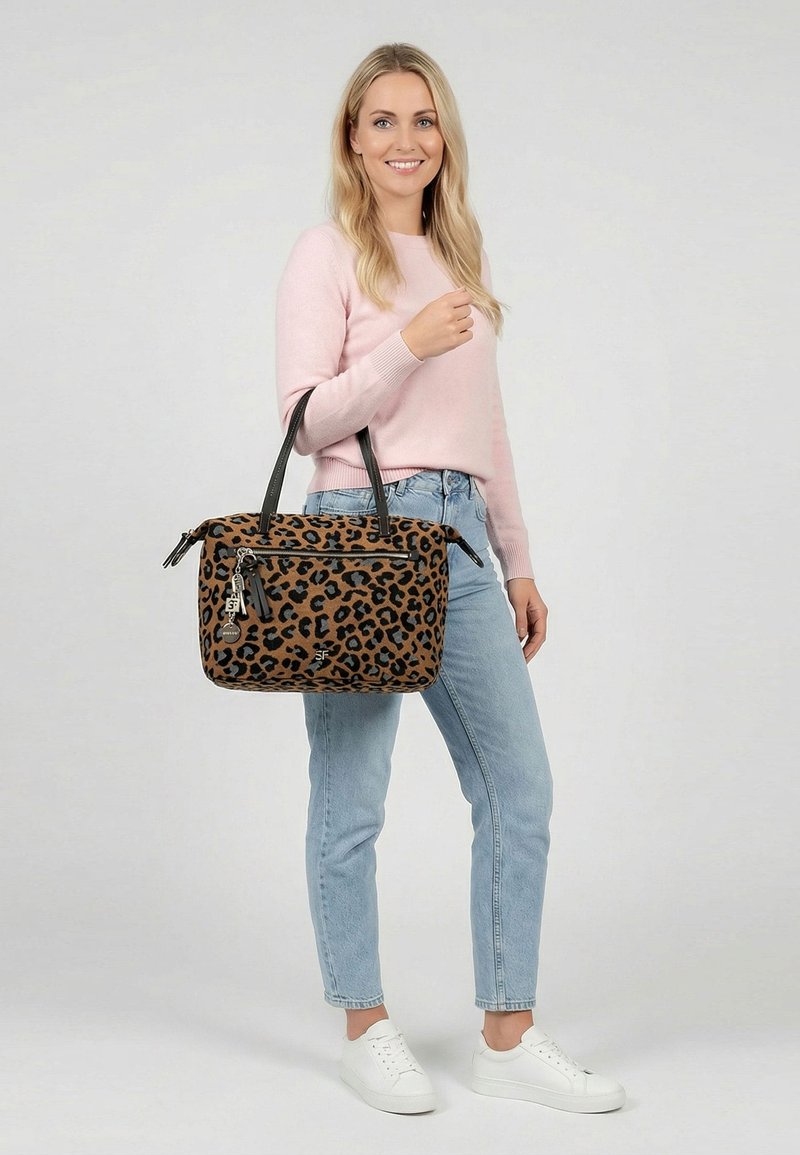 Femme portant un pull rose clair et un jean tenant un sac à main imprimé léopard marron avec des sangles noires et des détails zippés.