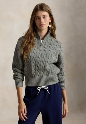 Junge Frau trägt einen grauen Zopfmuster-Pullover mit Viertelreißverschluss und marineblaue Jogginghose mit weißem Kordelzug vor neutralem Hintergrund.