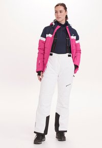 Roze geïsoleerde jas met marineblauwe accenten en witte panelen; gecombineerd met witte skibroeken met zwarte afwerking en verstelbare banden.