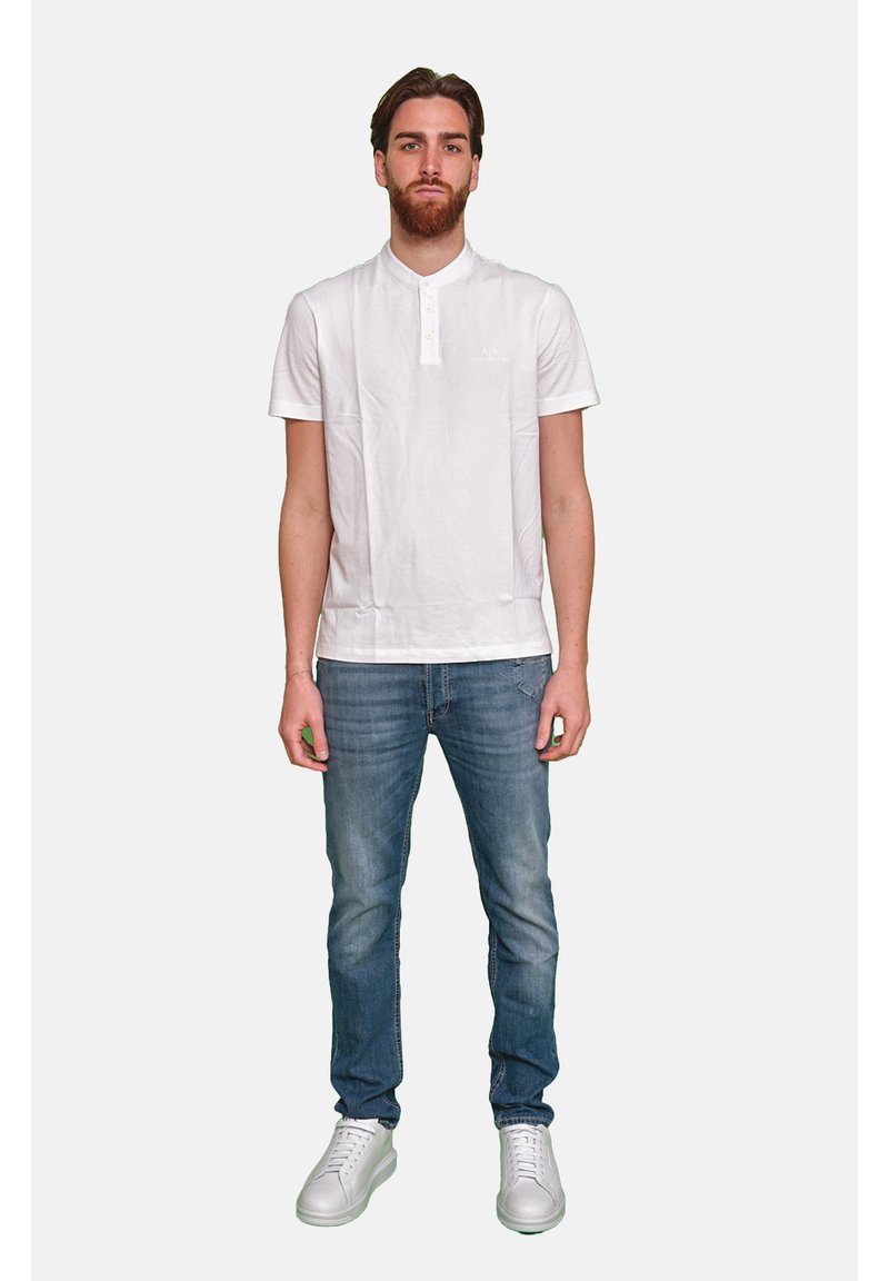 Polo shirt bianco a maniche corte con collo a bottoni, abbinato a jeans blu in denim e sneakers bianche, su uno sfondo semplice.
