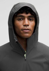 Ung man med lockigt hår iförd en mörkgrå hoodie, vänd framåt med ett neutralt uttryck mot en enhetlig grå bakgrund.