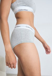 Sivi pamučni donji veš s visokim pojasom s bijelim logom Calvin Klein. Glatka tekstura i prilagođen dizajn, prikazan bočni pogled.
