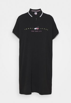 Černé polo šaty z bavlny s klasickým límečkem, krátkými rukávy a bílým lemováním. Nápis "TOMMY JEANS NEW YORK CITY" je potištěn barevnými písmeny.