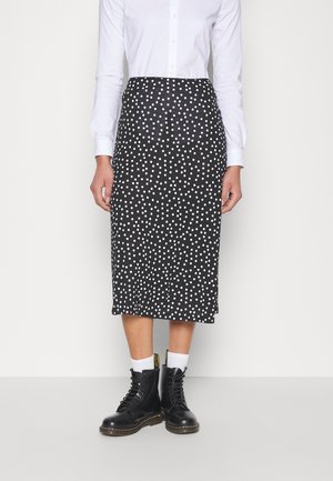 Person iført en sort midi-nederdel med hvide polkaprikker, hvid skjorte med knapper, hvide sokker og sorte snørestøvler, stående på en ensfarvet baggrund.