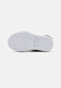 A Lacoste tornacipő talpa látható, amely egy fehér, rugalmas gumiból készült aljzatot mutat be, zigzag mintázattal és márkázott logó részlettel.