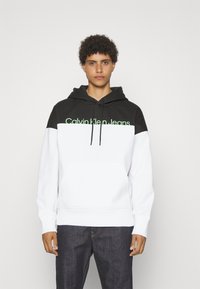 Mikina s kapucňou v čiernej a bielej farbe, s klokankou, rebrovanými manžetami a logom "Calvin Klein Jeans" v zelenej farbe na hrudi.