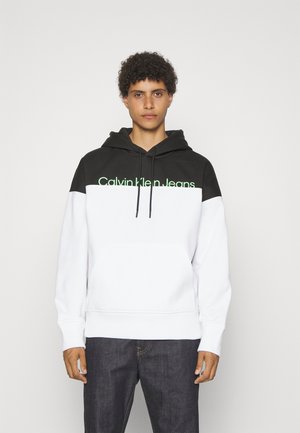Czarna i biała bluza z kapturem, z kieszenią kangurzą, ściągaczami przy rękawach oraz zielonym logiem "Calvin Klein Jeans" na piersi.