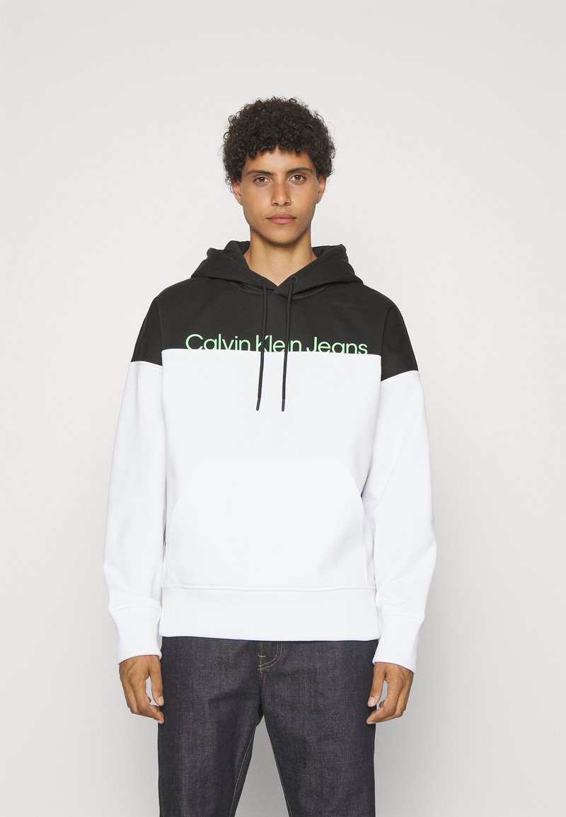 Mikina s kapucňou v čiernej a bielej farbe, s klokankou, rebrovanými manžetami a logom "Calvin Klein Jeans" v zelenej farbe na hrudi.