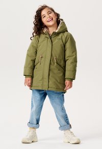 Olivenfarvet, hætteparka med snor i taljen og lommer. Foret med mønstret stof. Sammen med opsmøget blå jeans og lyse sneakers.