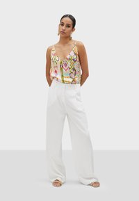 Top senza maniche a fantasia colorata con scollatura a V profonda, abbinato a pantaloni bianchi a vita alta e a ghette piatte con dettagli dorati.