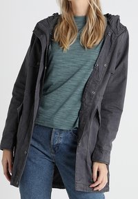 Šedá kapucínová bunda s předním zipem a cvoky, s velkými kapsami a volným střihem, nošená přes tyrkysové tričko a džíny.