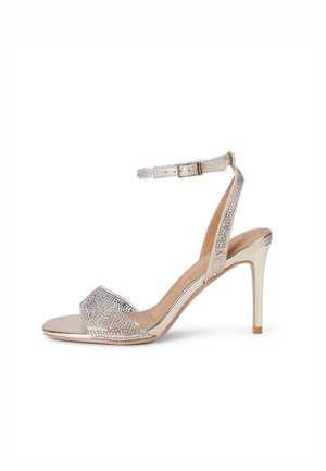 Sandale à talon haut argentée avec bride à la cheville et bande sur les orteils, toutes deux ornées de petits strass, sur fond blanc.