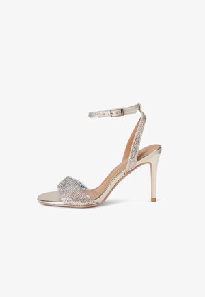 Sandale à talon haut argentée avec bride à la cheville et bande sur les orteils, toutes deux ornées de petits strass, sur fond blanc.