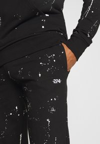 Main droite dans la poche d'un pantalon de survêtement noir avec un motif d'éclaboussures de peinture blanche et un détail brodé "274" en blanc près de la poche.