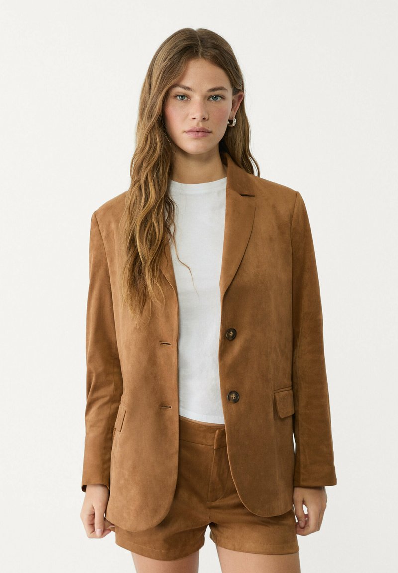 Brauner Wildleder-Blazer mit taillierter Passform, zwei Knöpfen und Seitentaschen, kombiniert mit einem weißen Hemd und passenden braunen Shorts.