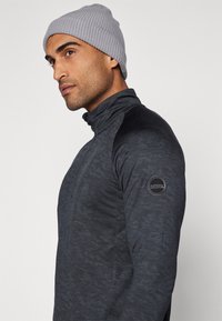 Suéter ajustado de color gris oscuro con cuello alto y cremallera, tejido texturizado, paneles en negro en los hombros y parche con logo en la manga izquierda.