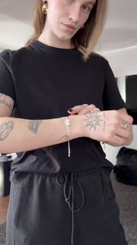 Sidabrinė apyrankė su žvaigždės pakabuku, turinti subtilią grandinėlę. Ant dėvėtojo rankų matosi tatuiruotės, jis apsirengęs juodą marškinėlį.