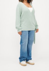 Mintgrön texturerad cardigan med V-ringning och knytdetalj, kombinerad med ljusblå denimjeans och vita sneakers med tanfärgade sålar.