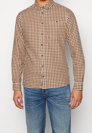 Homme portant une chemise à carreaux marron et blanc à manches longues boutonnée, avec un jean bleu, debout devant un fond clair uni.