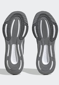 Suole di scarpe sportive grigie con gomma testurizzata, caratterizzate da un motivo a onda e accenti bianchi. Il design include scanalature allungate per una migliore aderenza.