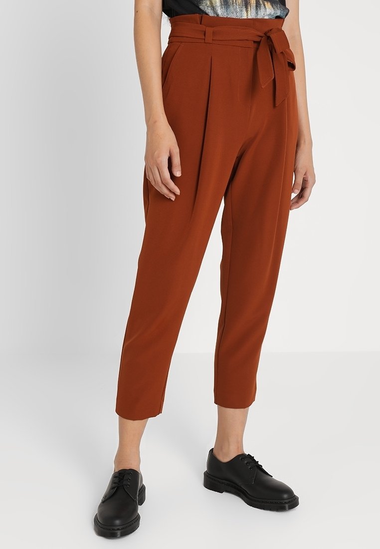 Pantalon fuselé taille haute couleur rouille avec plis et ceinture en tissu nouée à la taille, porté avec des chaussures noires à lacets.