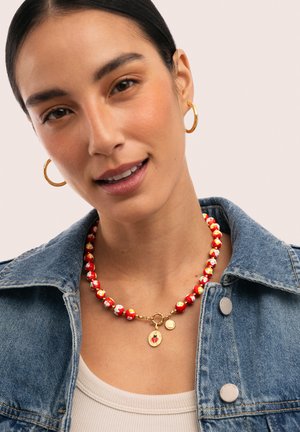 Frau mit goldenen Creolen, einer rot-weißen Perlenkette mit Marienkäfer-Anhänger, einem weißen Top und einer blauen Jeansjacke.