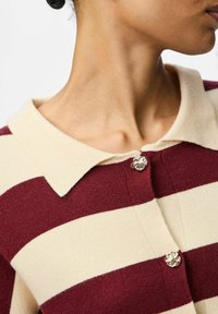 Karamellbrūns un krēmkrāsas svītrains kardigans ar apkakli un dekoratīvām sudraba lauva galvas pogām. Mīksts adījuma tekstūra. Tuva kakla izgriezuma un auduma attēls.