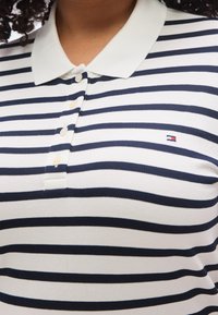 Ριγέ πόλο μπλουζάκι σε λευκό με ναυτικό μπλε οριζόντιες ρίγες, με κλασικό γιακά, κουμπότρυπα και μικρό λογότυπο της μάρκας στο στήθος.