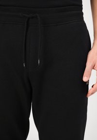 Černé tepláky vyrobené z měkké tkaniny s elastickým pasem, šňůrkou na stažení a bočními kapsami. Na jedné noze je decentní logo.